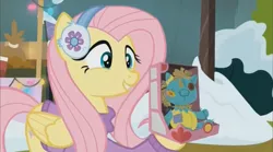 Size: 1352x754 | Tagged: safe, derpibooru import, screencap, fluttershy, holly the hearths warmer doll, pegasus, pony, best gift ever, clothes, cute, earmuffs, female, happy, mare, shyabetes, smiling, solo, sweater, winter