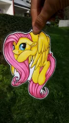 Size: 670x1192 | Tagged: safe, artist:julunis14, derpibooru import, fluttershy, pegasus, pony, female, floppy ears, hand, holding a pony, irl, laminated, mare, neck hold, photo, scruff, solo, three quarter view, traditional art, wings