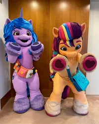 Size: 1080x1350 | Tagged: safe, derpibooru import, official, izzy moonbow, sunny starscout, anthro, earth pony, human, pony, unicorn, 2022, bag, belt, cutie mark, fursuit, g5, image, irl, irl human, jpeg, mascot, mascot costume, outstretched arms, photo, ponysuit, raised hoof, smiling, wide eyes, wide hips